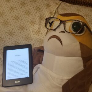 A Porg stuffed animal, laying in bed, wearing glasses, reading a book on a Kindle