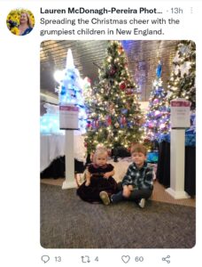 Screenshot of a Tweet that reads "Spreading the Christmas cheer with the grumpiest children in New England". Attached is a picture of two small children, sitting in front of a Christmas tree, looking annoyed.