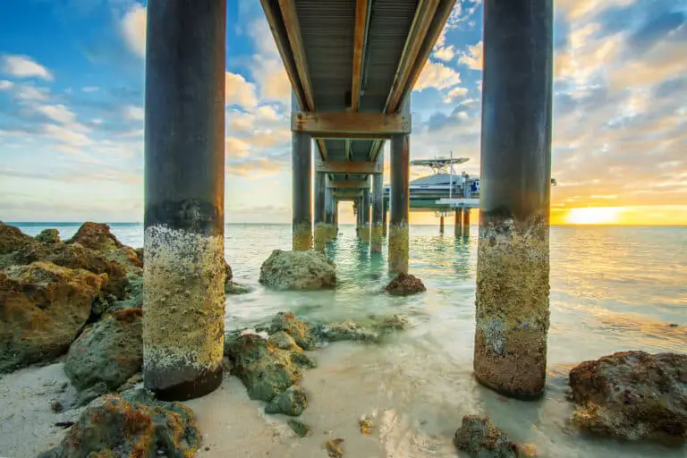 An image of a wooden pier over a beach at sunset. The pier is weathered with barnacles attached to the legs. The shore is sandy with large rocks. The sunset makes the sky blue on the left side, and yellow on the right.