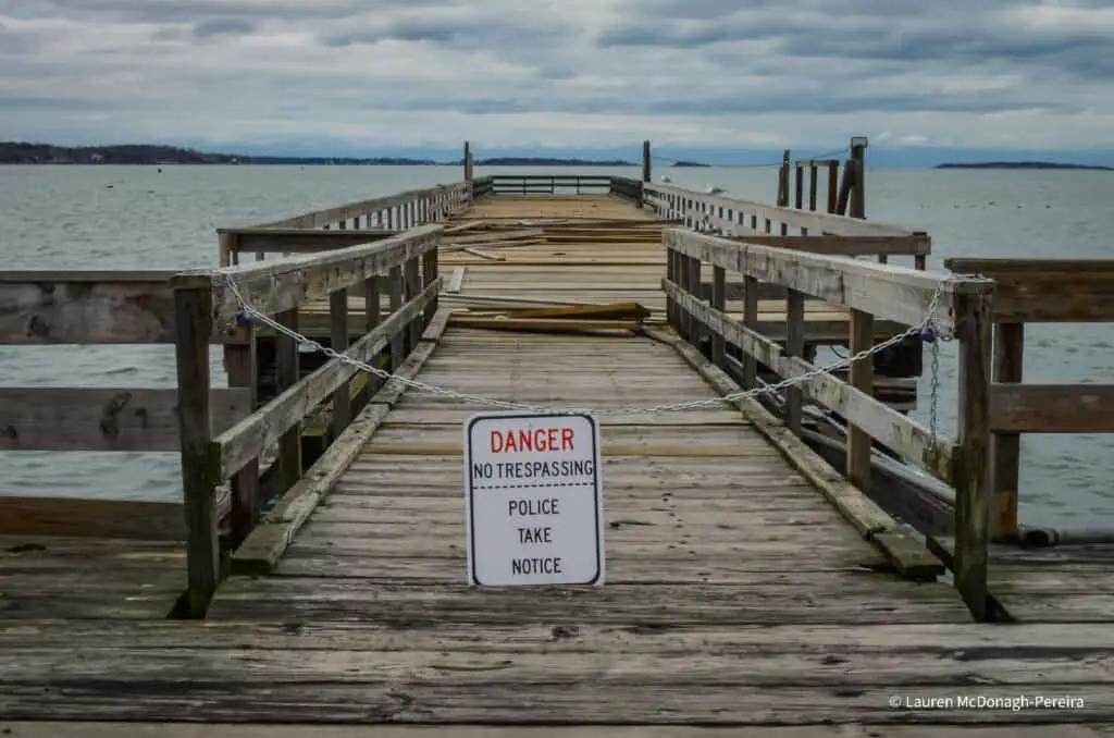 An image of a damaged wooden pier leading to the calm sea. A chain block the entrance to the pier. A sign on the chain reads "Danger - No trespassing. Police take notice."