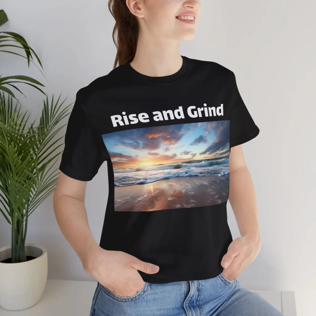 A woman wearing a black tshirt that says Rise and Grind with the sun rising over the ocean. 