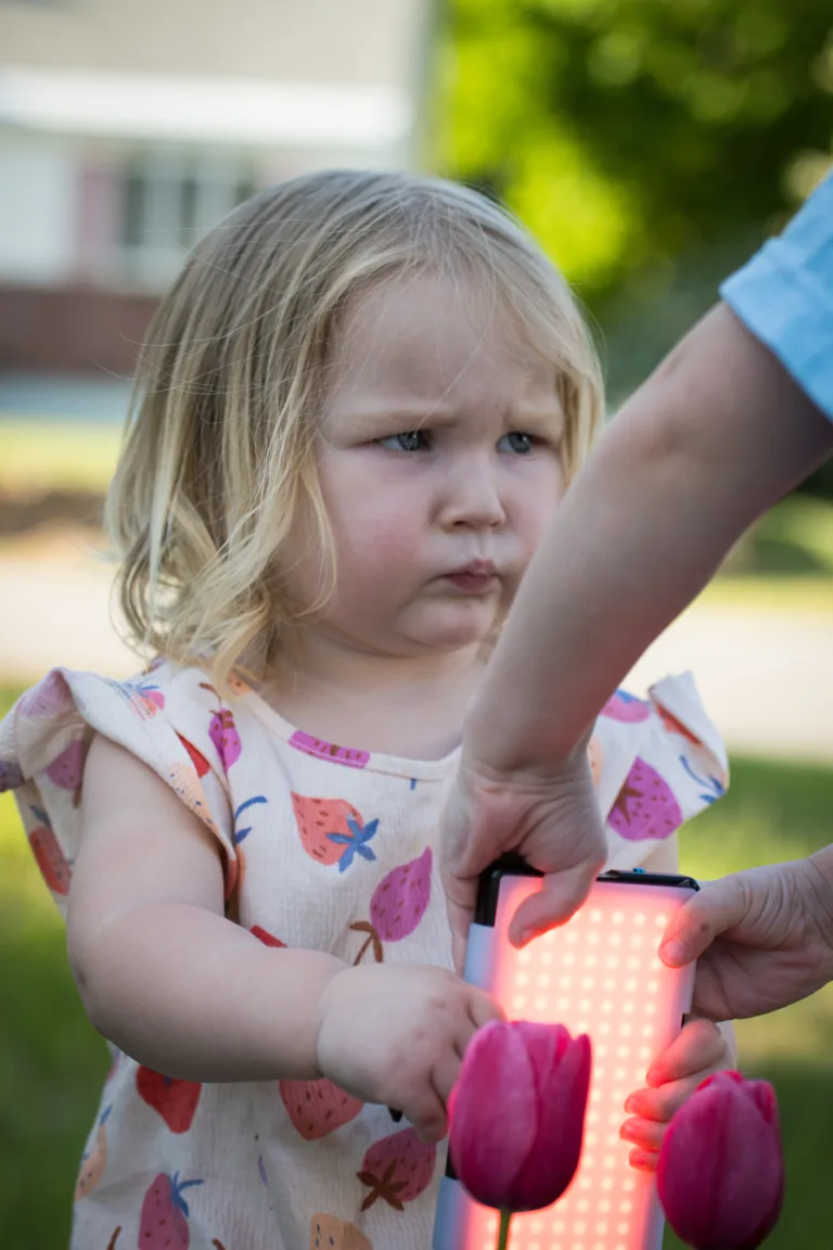 A one year-old girl holds a lume cube and wrinkles her nose with annoyance as her older brother tries to steal it.